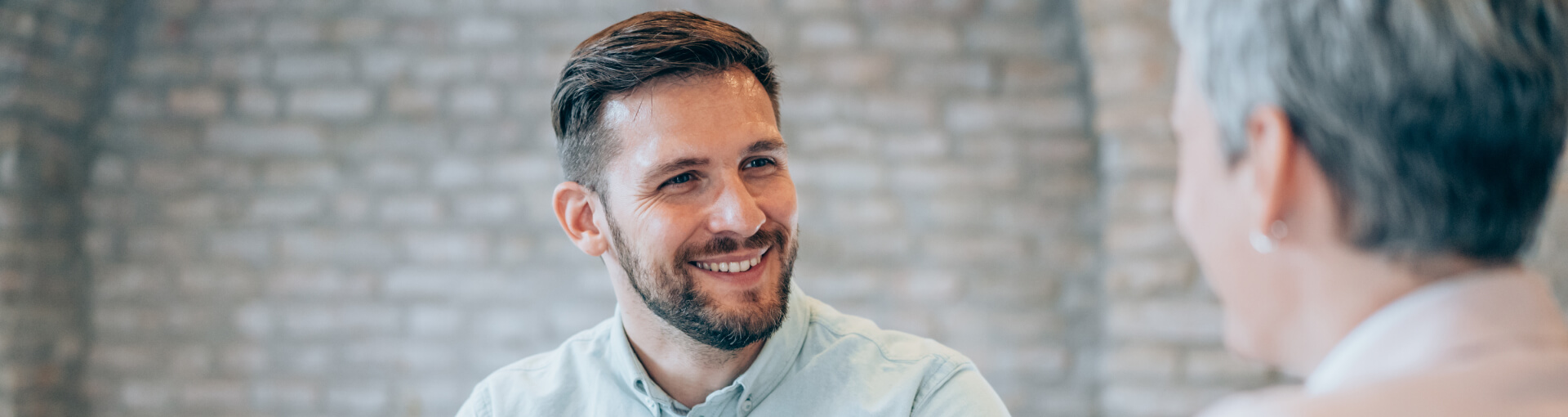 Smiling man with a beard in a light shirt talking with a woman in front of a brick wall