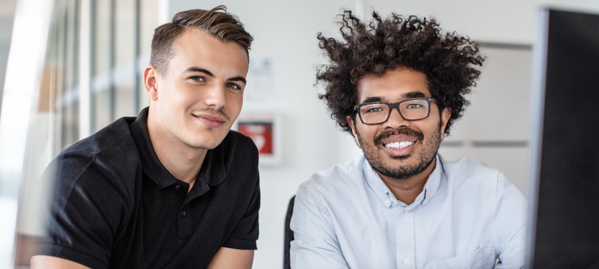 Two smiling students, one in a black polo shirt, one with curly hair and glasses, in an office