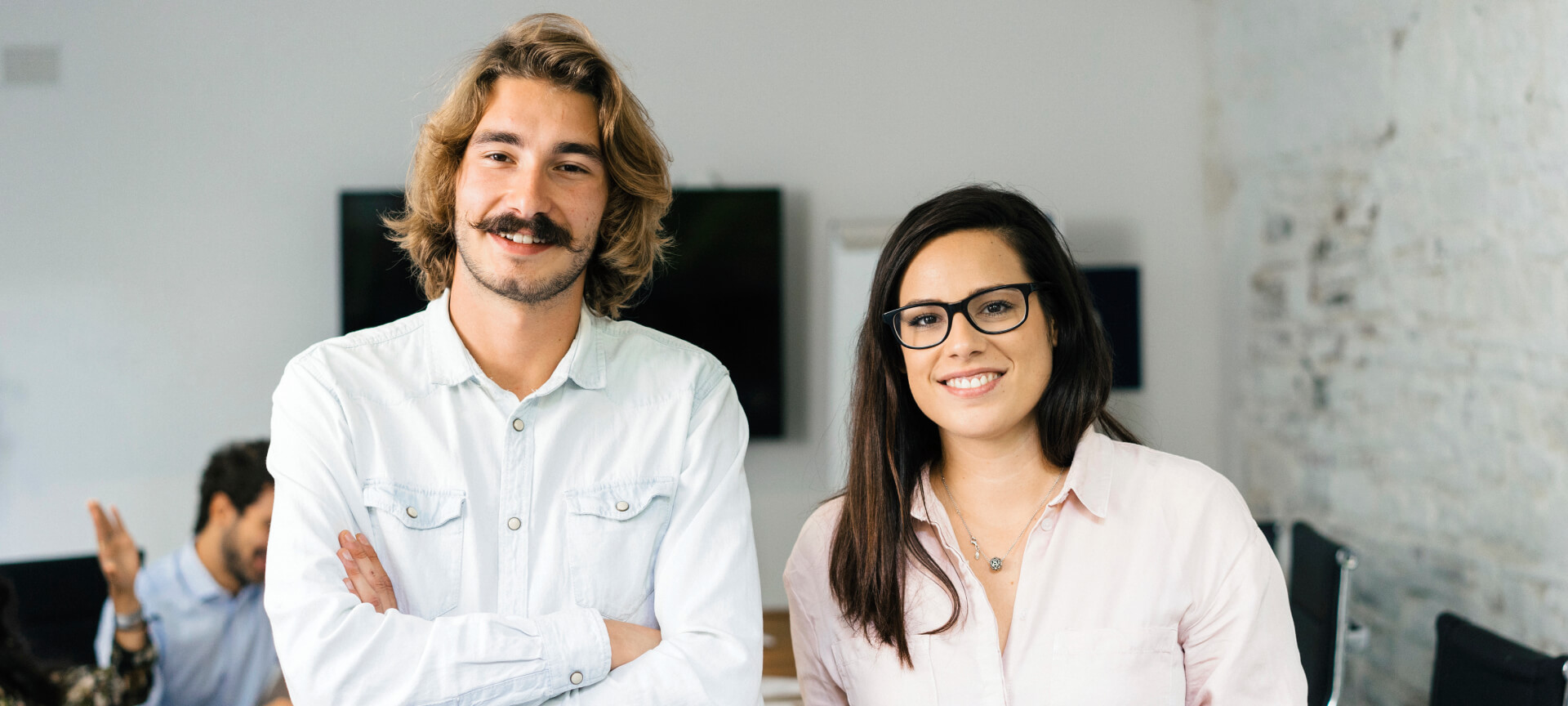 Two smiling young professionals, he with a moustache, she with glasses, standing in an office