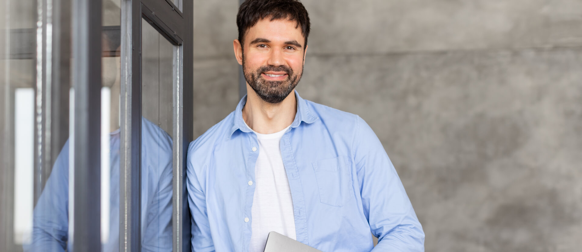 Smiling expert with a beard and light blue shirt leaning on a glass door with a laptop
