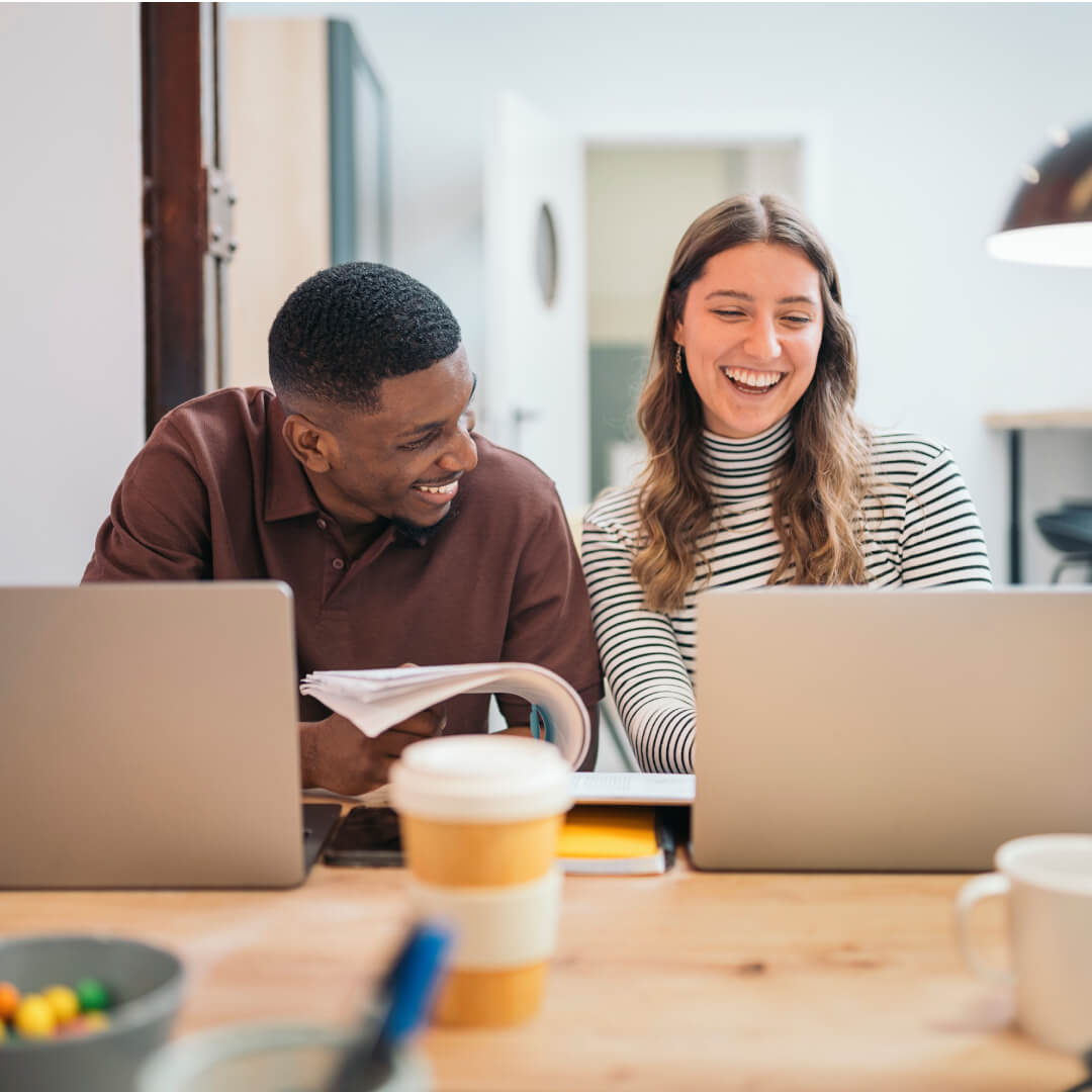 Two colleagues laughing together at a desk with laptops and a coffee cup