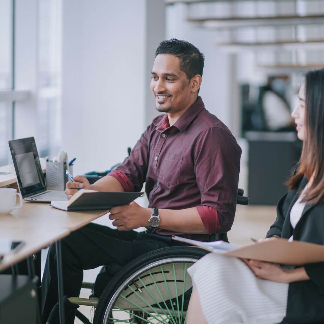 Smiling man in a wheelchair holding a notebook, sitting next to a female colleague at an office desk