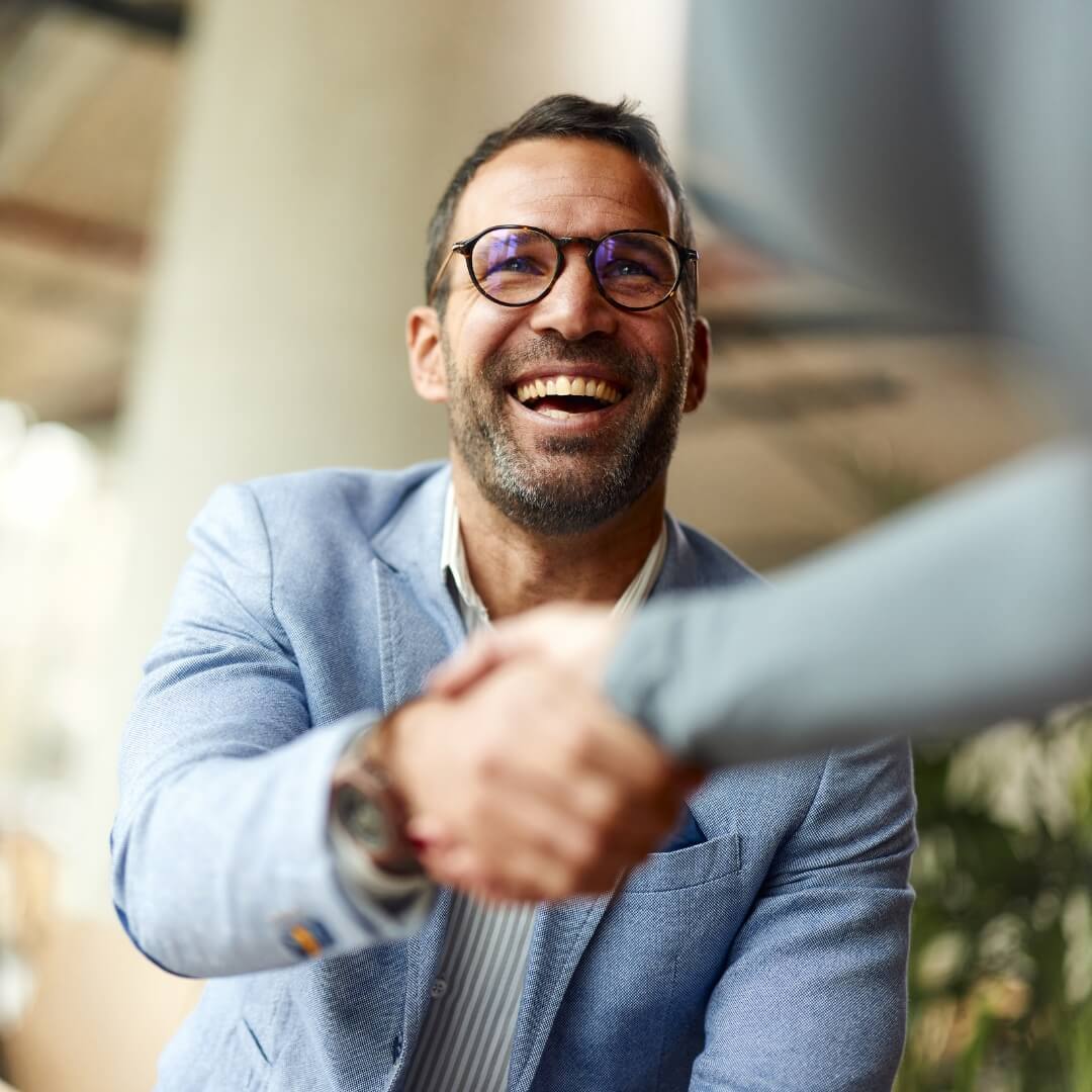 Laughing man with glasses and a light blue blazer shaking someone's hand in greeting
