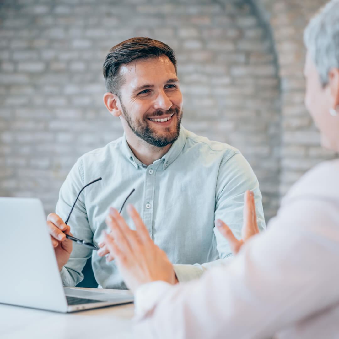 Smiling man holding his glasses, talking with a female colleague at a desk