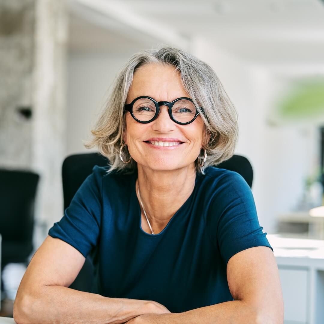 Smiling woman with gray hair and round glasses sitting in a bright office