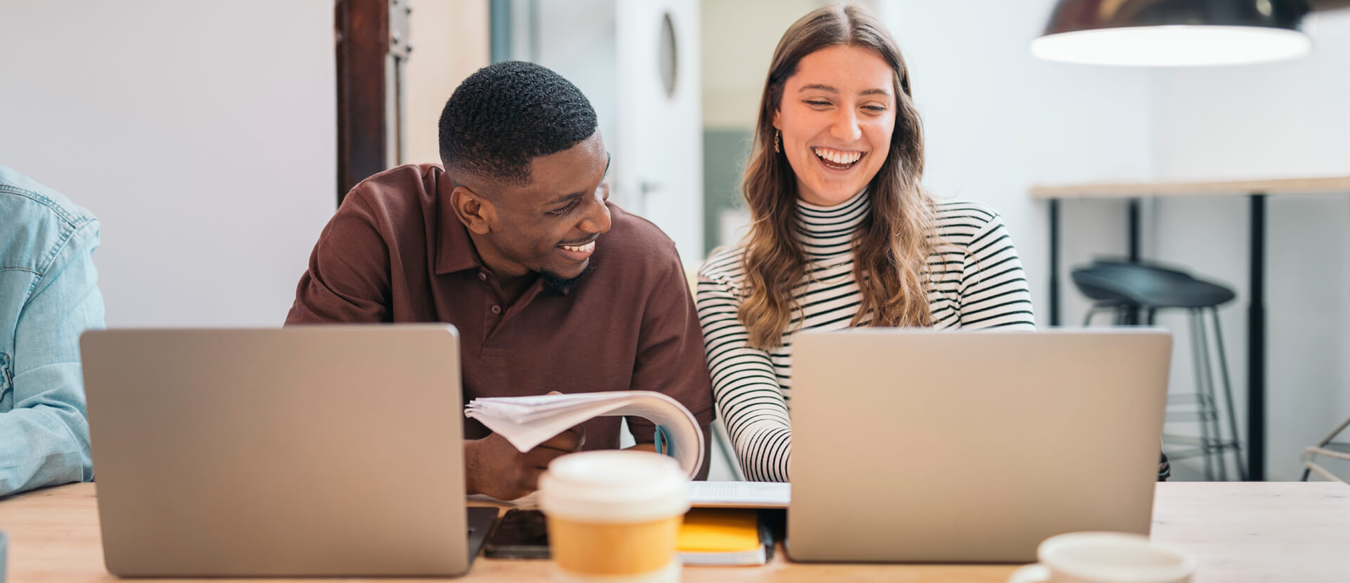 Kollege und Kollegin lachen gemeinsam am Schreibtisch mit Laptops und Kaffeebecher