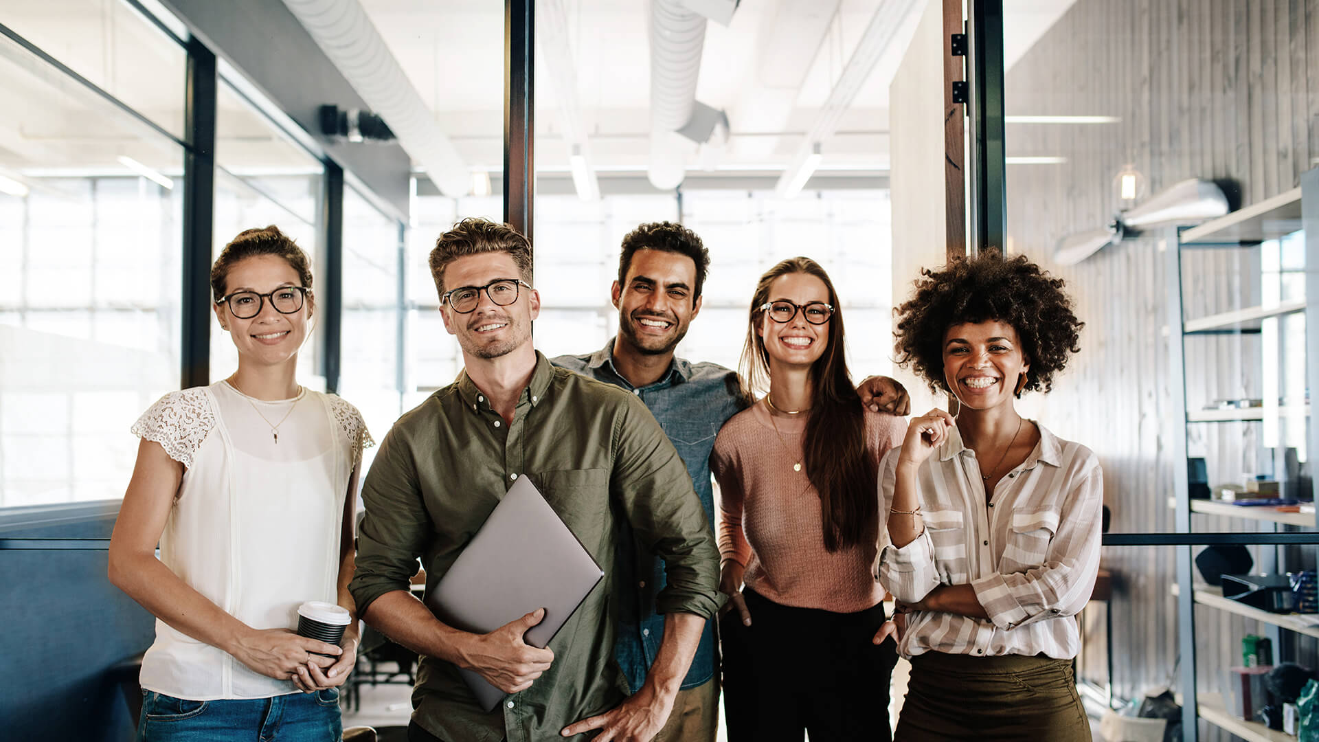 Fünfköpfiges Team steht lachend und gut gelaunt in einem modernen Loft-Büro