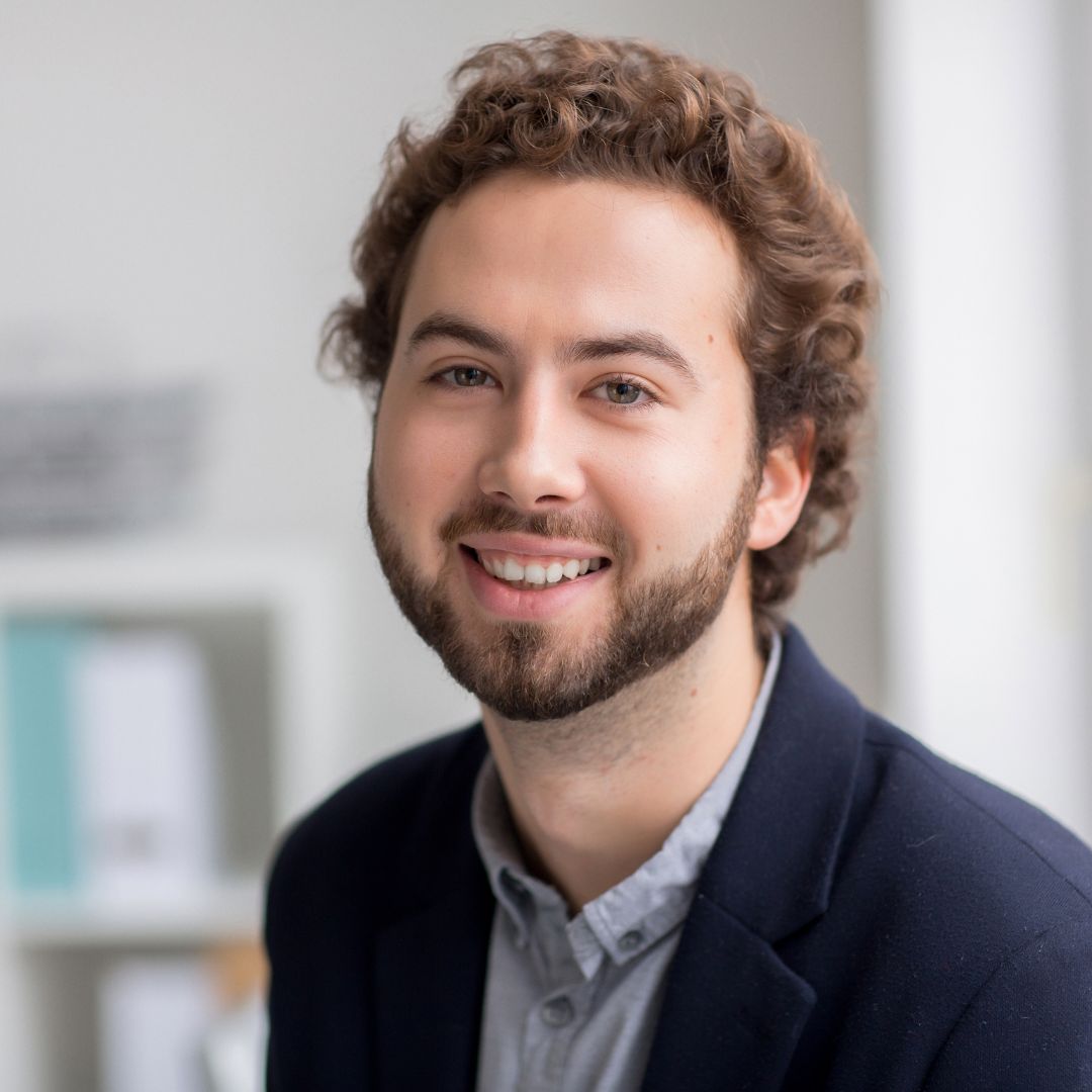 Lächelnder Student mit Locken und Bart im dunkelblauen Sakko in einem hellen Büro